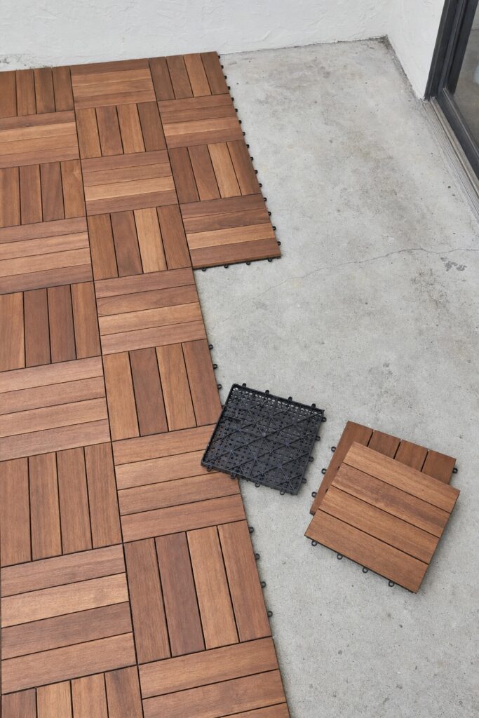 Interlocking wood deck tiles half laid over a bare concrete apartment patio slab showing the drainage grid underneath
