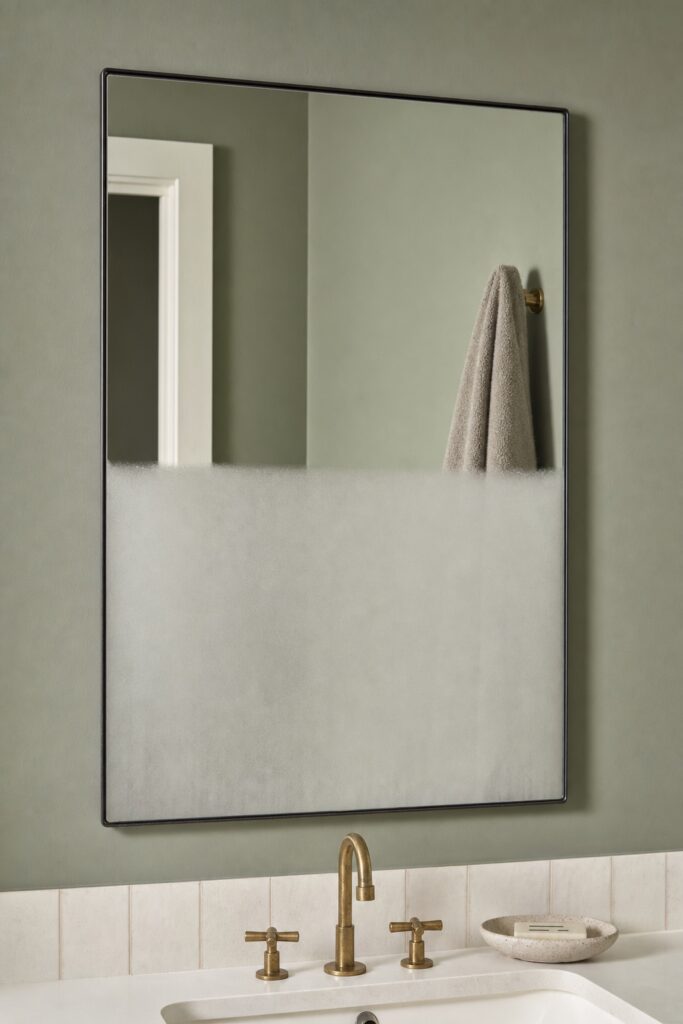 A bathroom mirror half cleared of fog after a shower, with the clear part reflecting the room