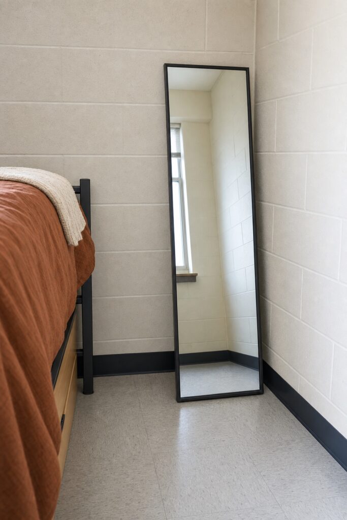 Full-length mirror leaning against the wall in the corner of a dorm room out of the walking path