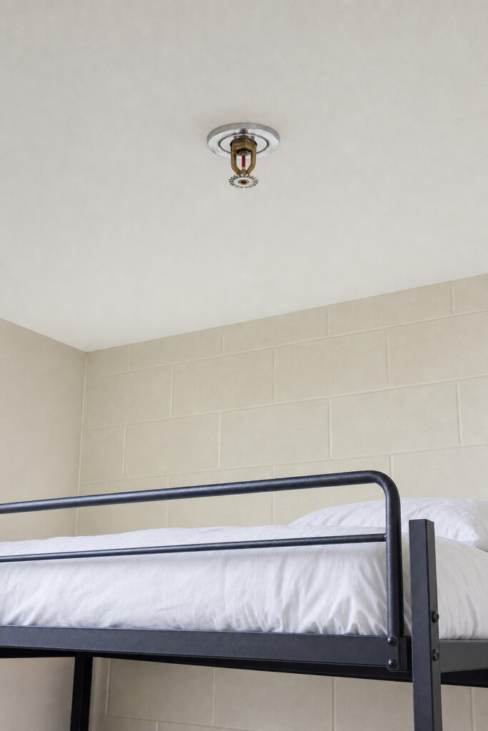 A sprinkler head hanging below the ceiling directly above a lofted dorm bed, with clear air between it and the mattress