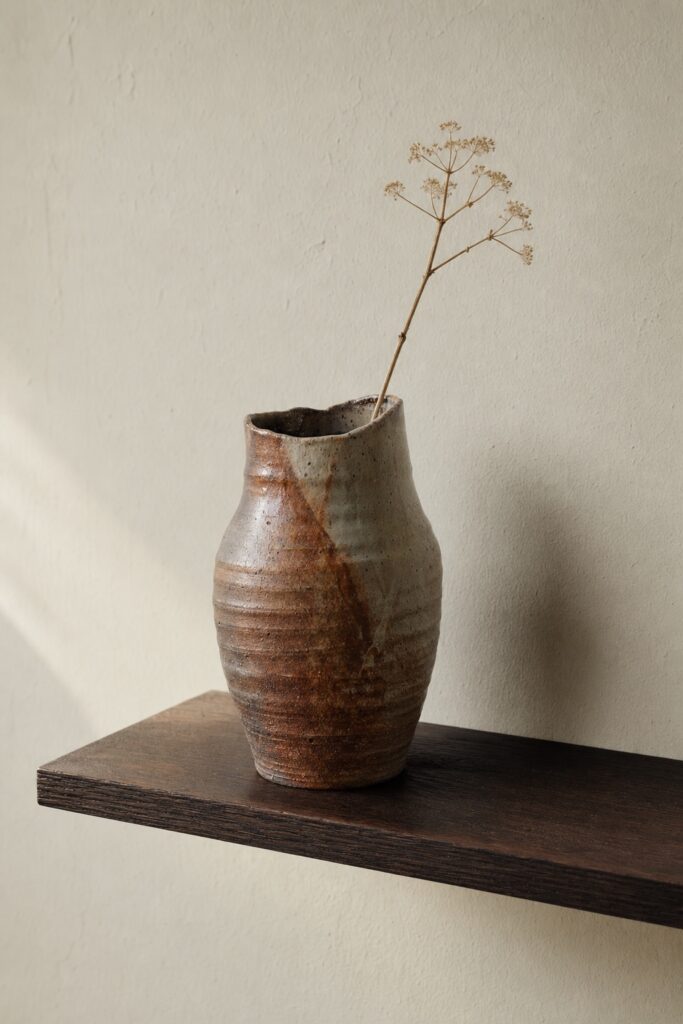 A single hand-thrown clay vase with a visibly uneven rim on a plain wooden shelf