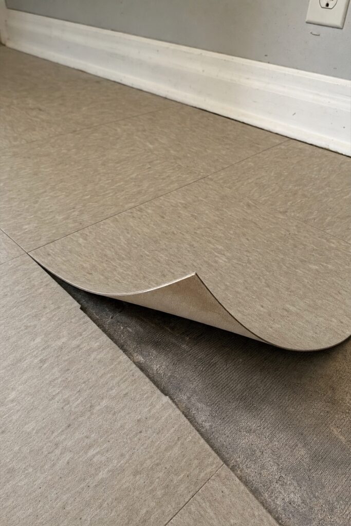 Corner of a peel and stick floor tile curling up away from a dusty textured floor