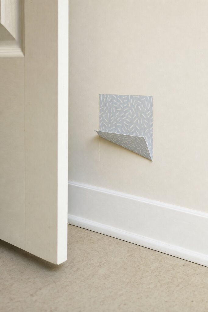 Small square adhesive test patch being peeled back low on a hidden wall showing clean undamaged paint underneath