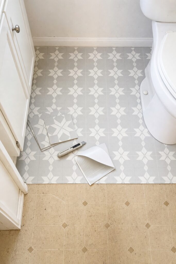Peel-and-stick vinyl tile being laid across a small bathroom floor with part of the old floor still visible