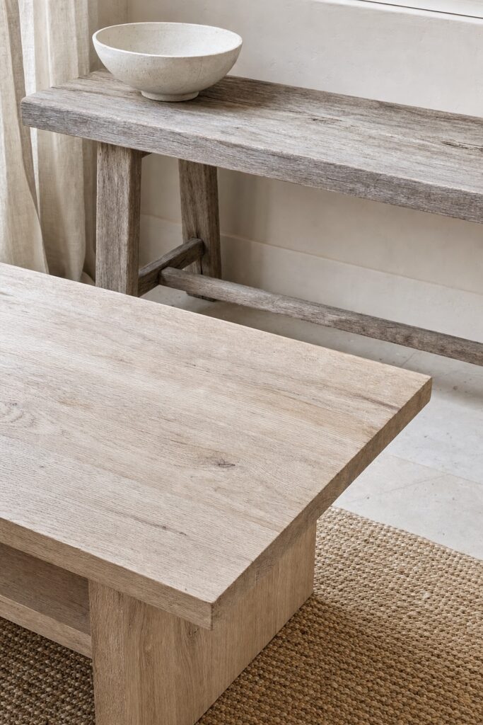 Pale oak coffee table and weathered gray teak bench with dry matte finishes in a light-filled coastal room