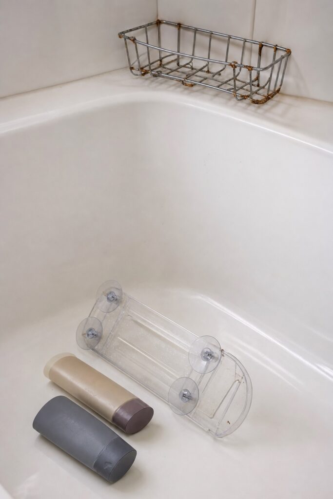 Fallen suction shelf with tipped bottles lying in a dry bathtub next to a chrome wire basket showing rust spots
